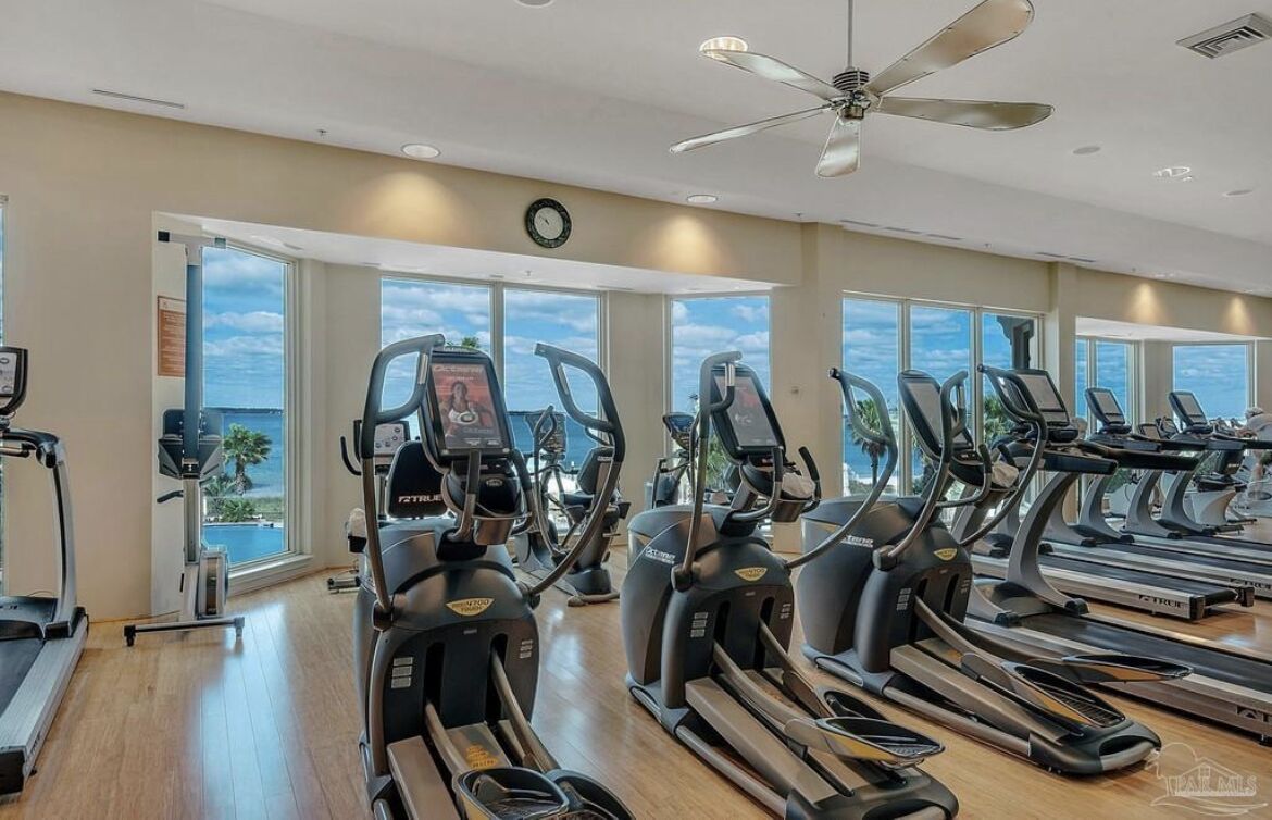 A gym with treadmills and elliptical machines, large windows with a view of the ocean, and a ceiling fan.