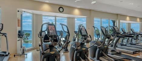 A gym with treadmills and elliptical machines, large windows with a view of the ocean, and a ceiling fan.