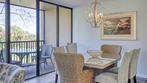 Un coin repas avec des chaises en osier et une table, un lustre et un tableau encadré représentant un oiseau accroché au mur.