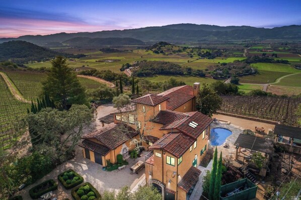 A villa with multiple buildings, a swimming pool, and vineyard fields in the background.