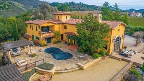Una casa grande de color amarillo con piscina, mobiliario exterior y vistas a las colinas circundantes.