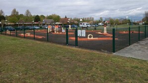 Cancha deportiva cercada con aro de baloncesto, rodeada por un estacionamiento con coches y un edificio al fondo.