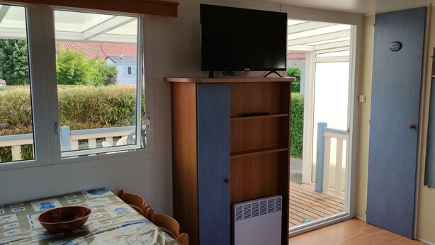 Una habitación con televisión sobre un mueble, una mesa con sillas y vistas a un jardín por un ventanal corredero.