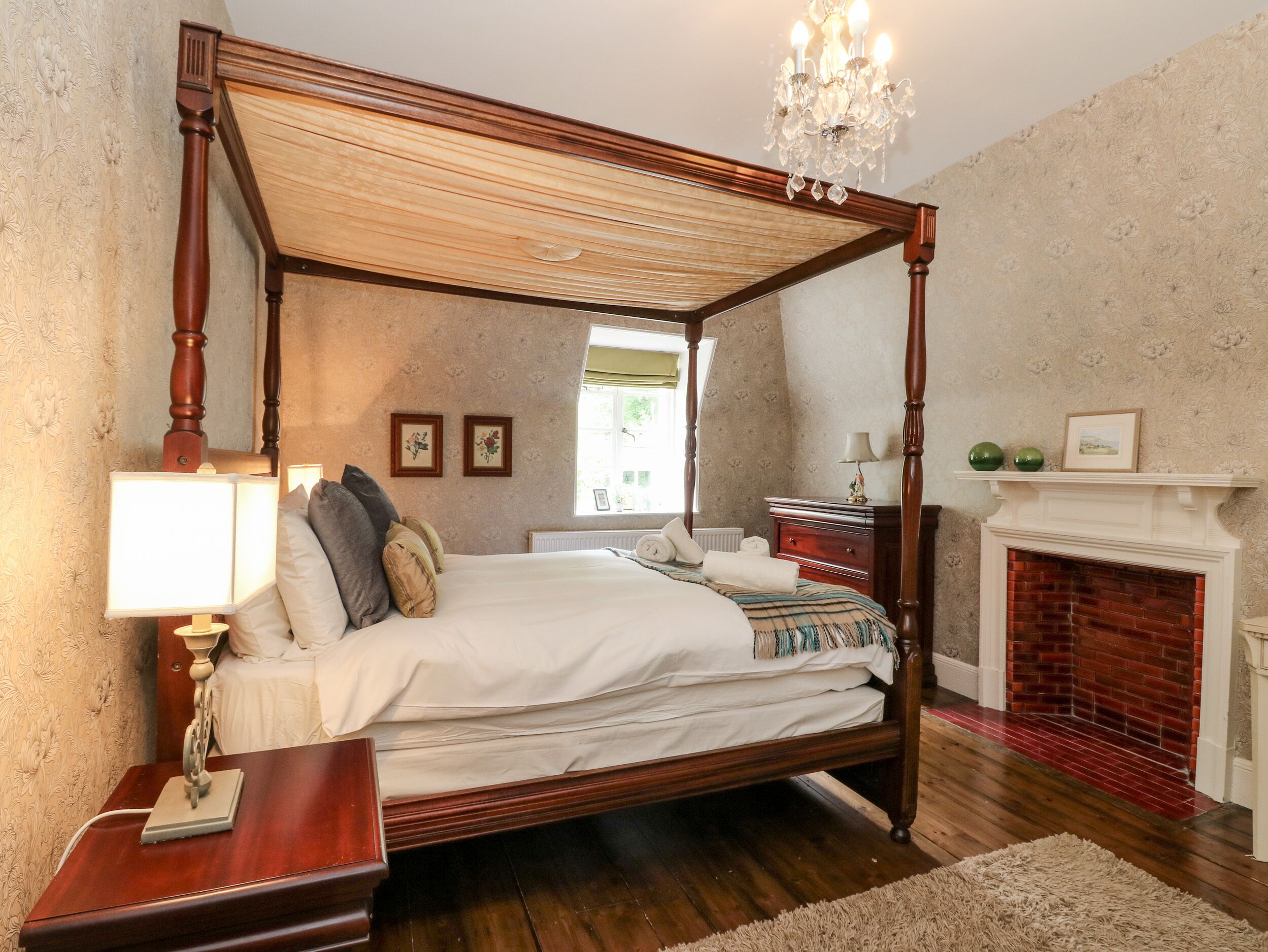 A four-poster bed with a canopy, a nightstand with a lamp, a fireplace, and a window with a view of greenery.