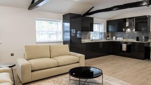A modern living room with a beige sofa, a black coffee table, and a kitchen with dark cabinets and a stone backsplash. - Holiday Lets Penrith – Derwent House (Appleby-In-Westmorland)