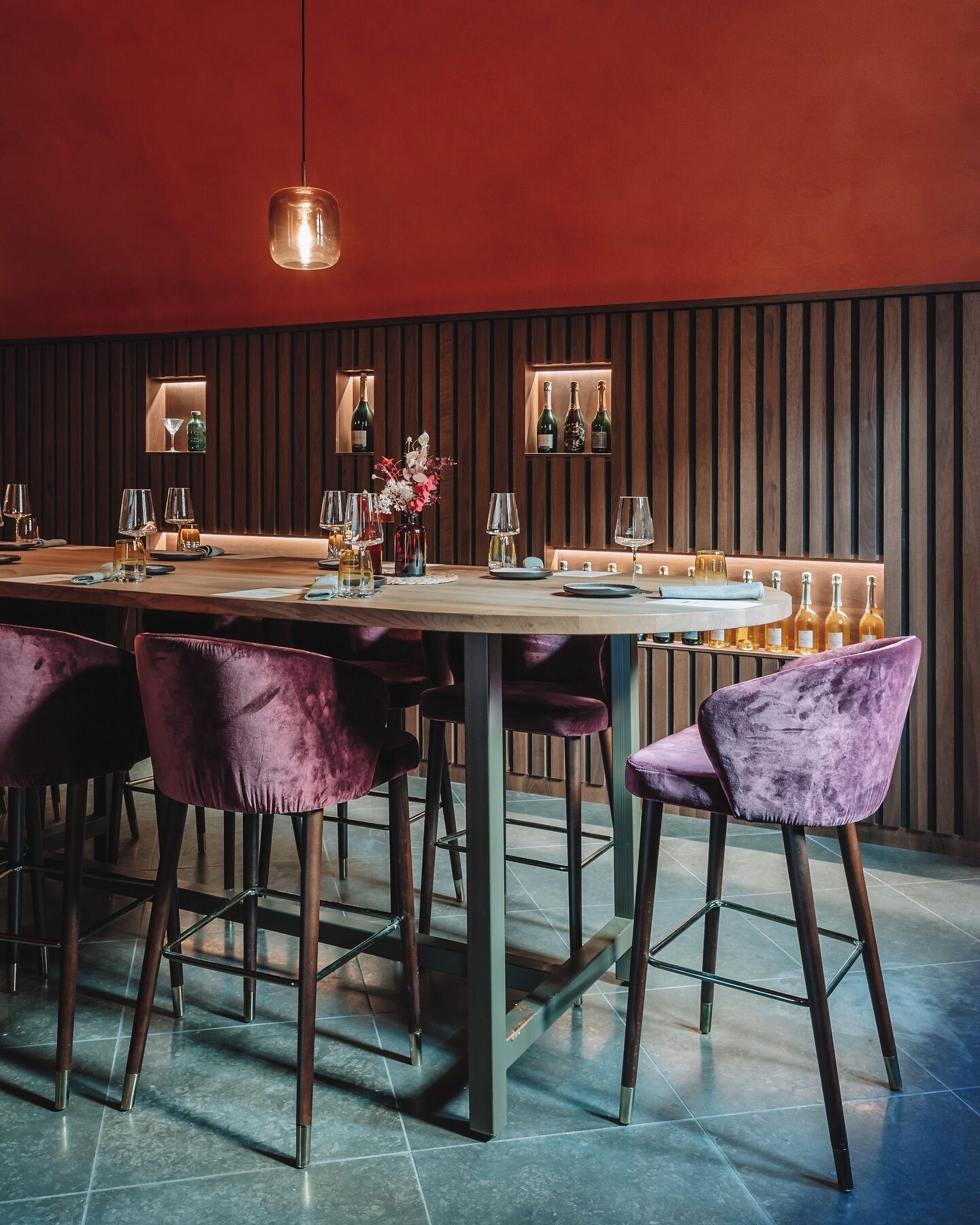Un espace bar avec une longue table en bois, des fauteuils en velours violet et des bouteilles de vin exposées sur des étagères.