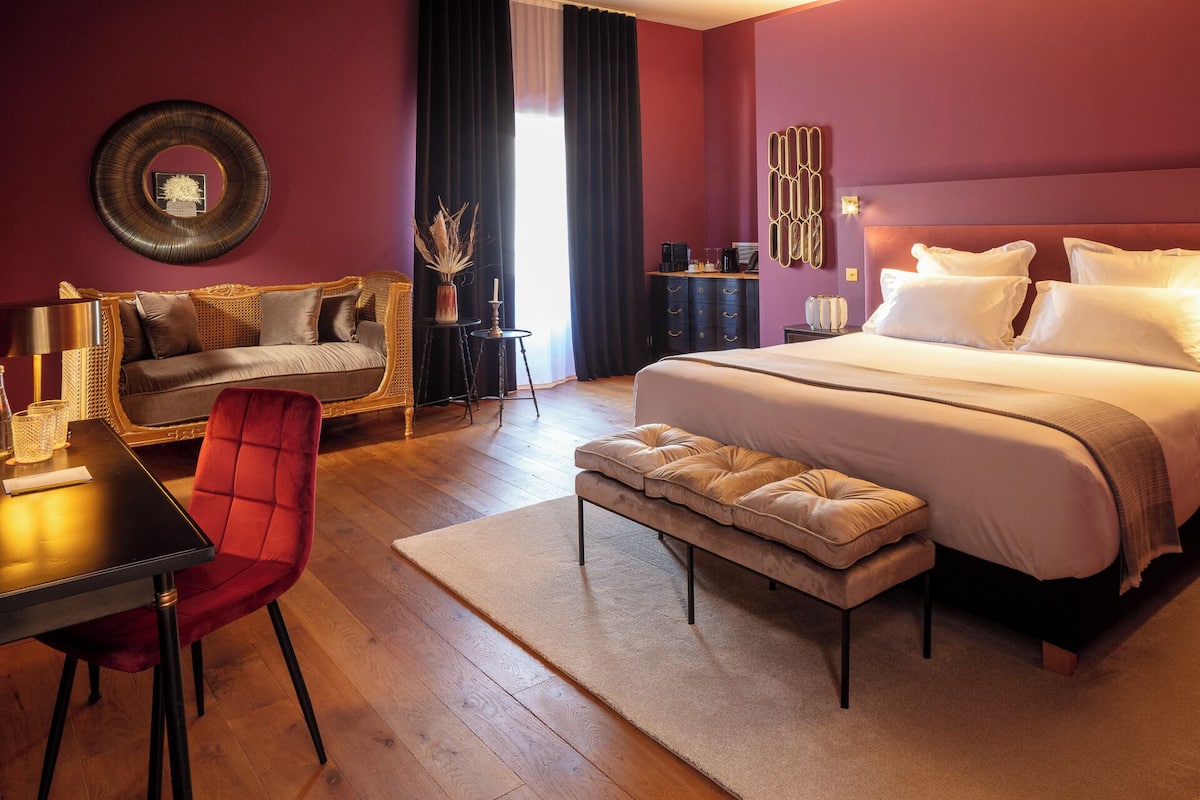 A modern hotel room with a large bed, a sofa, a red chair, a small table, and a decorative wall piece.