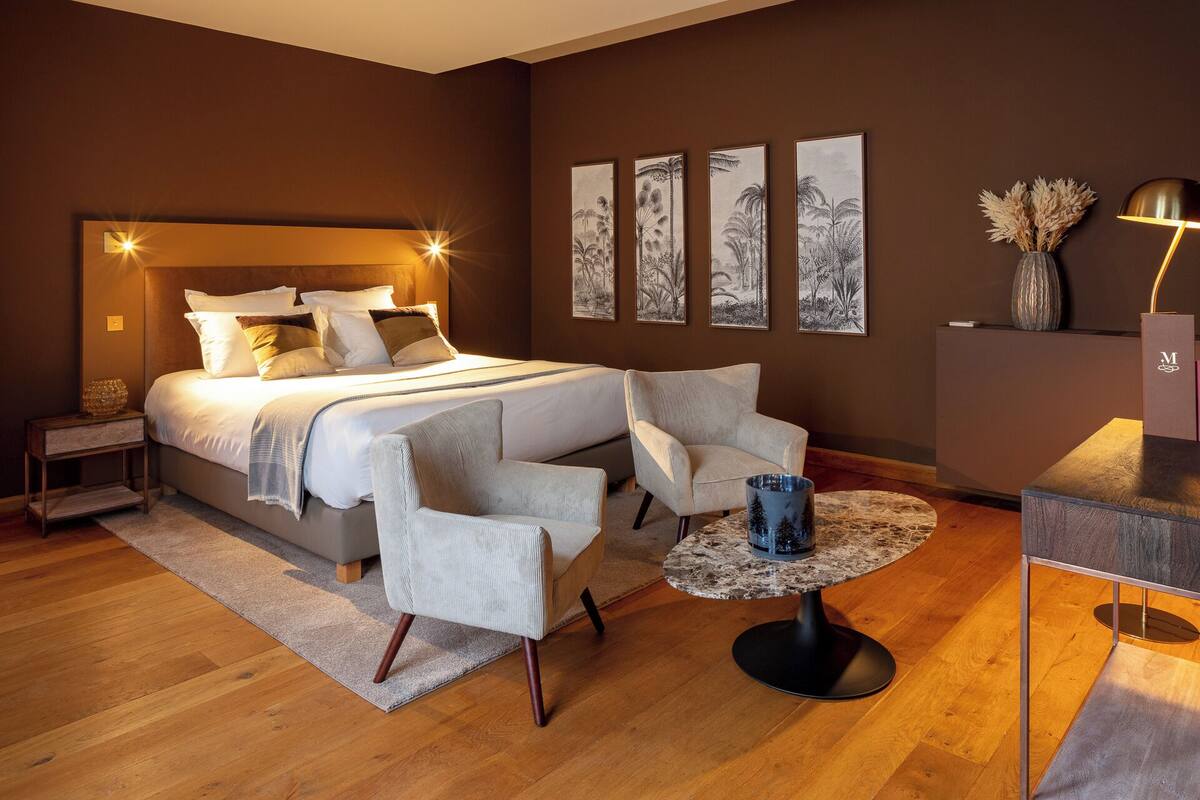 A bedroom with a large bed, two armchairs, a small round table, and a nightstand.