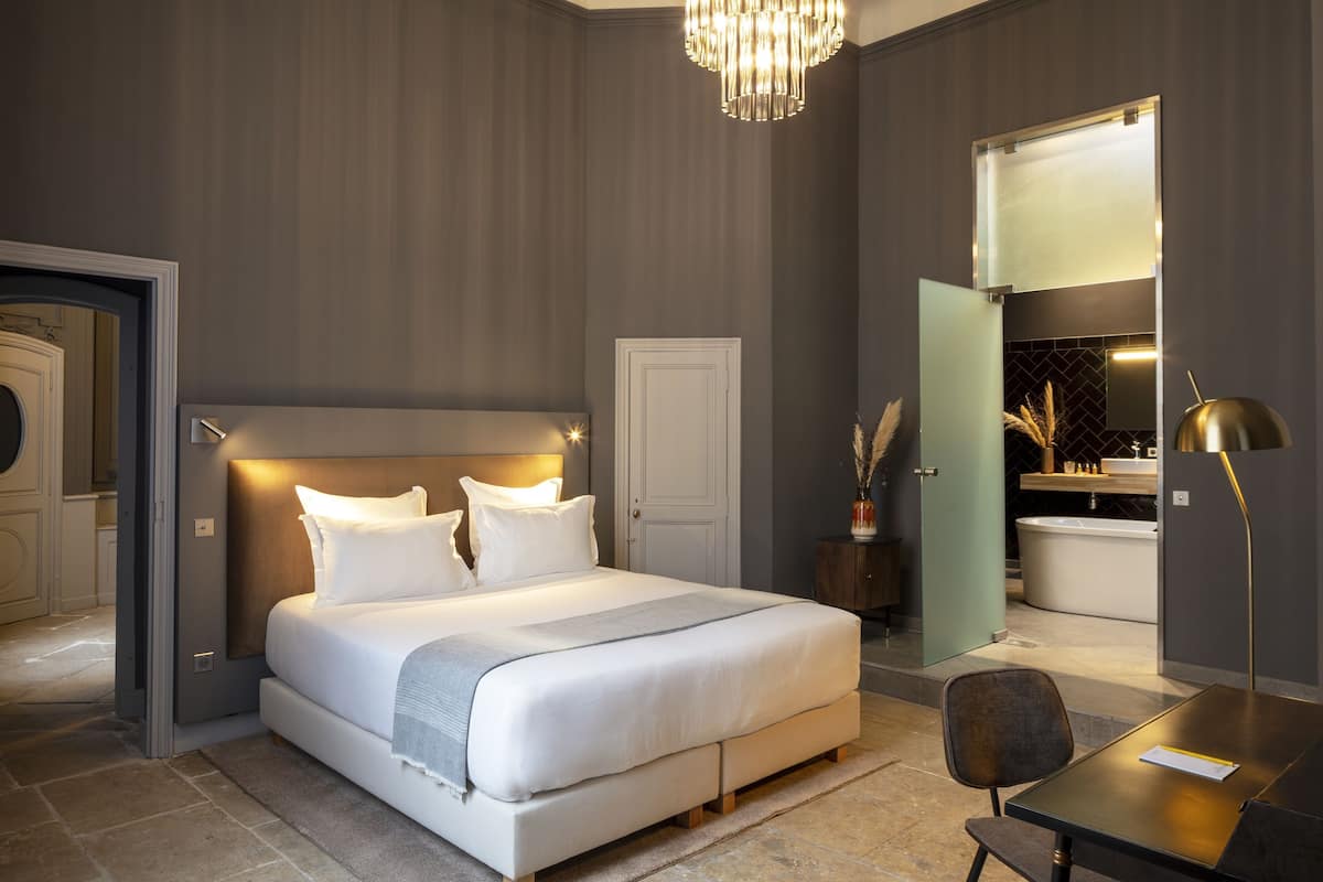 A modern bedroom with a large bed, bedside tables, a desk, and a bathroom visible through an open door.