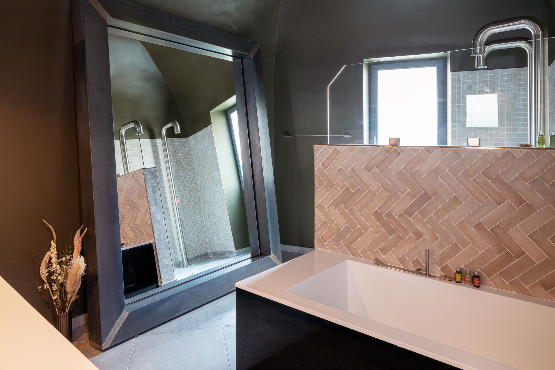 A modern bathroom with a large window, a herringbone-patterned wall, a white bathtub, and a mirror above the sink.