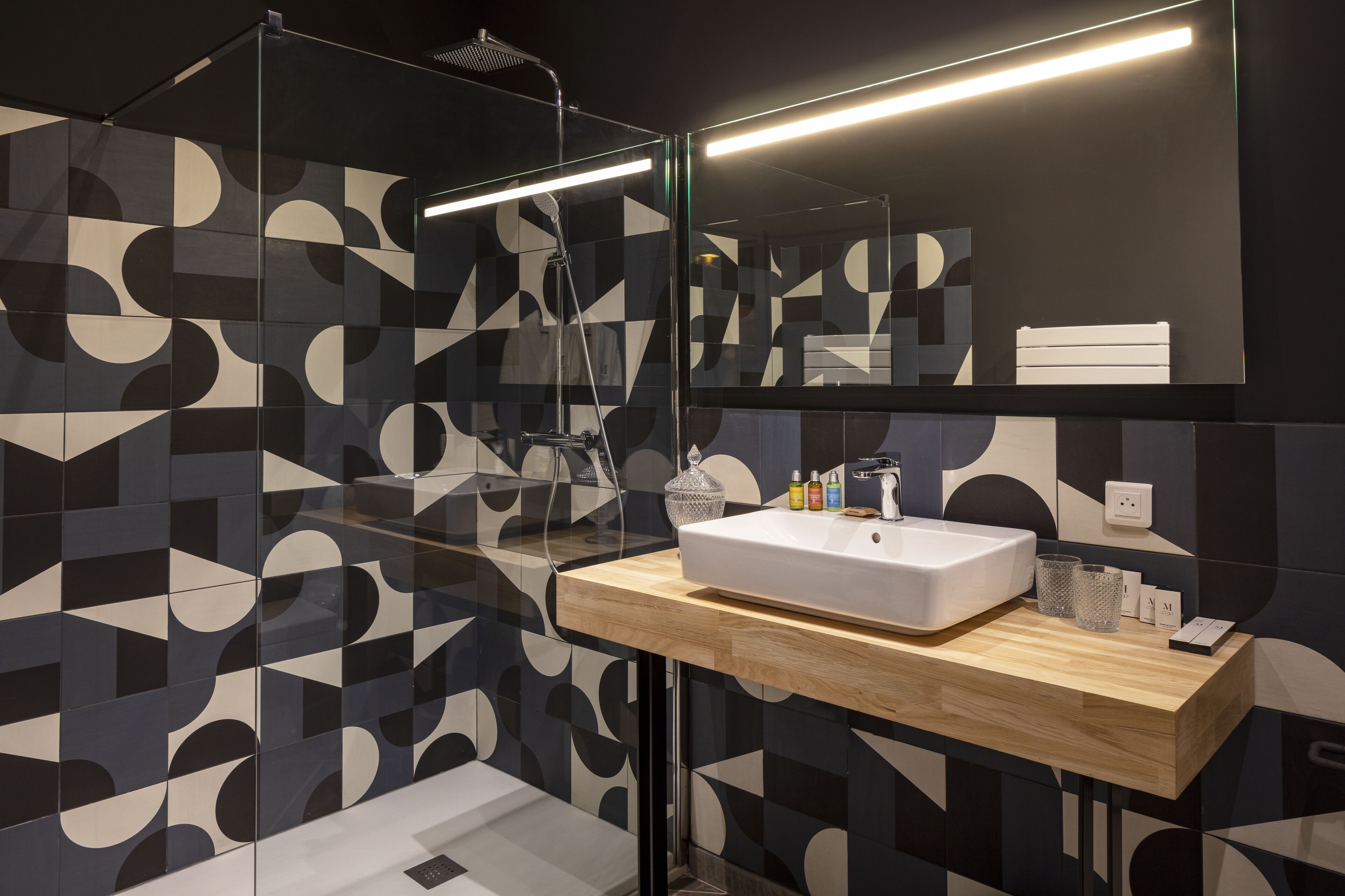 A modern bathroom with a geometric patterned wall, a wooden vanity, a white sink, and a glass shower enclosure.