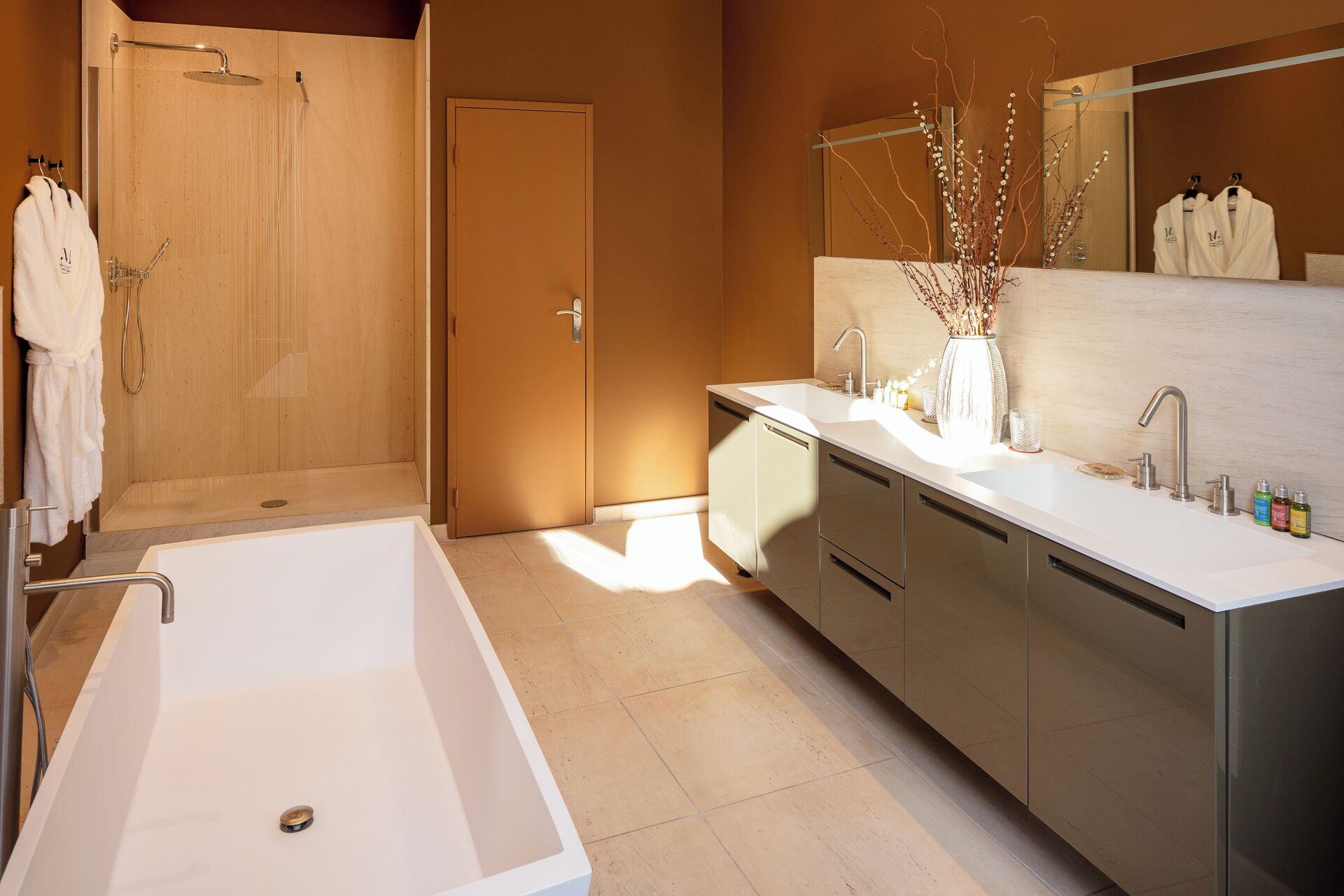 A modern bathroom with a large bathtub, a shower, a vanity with a mirror, and a vase with branches.