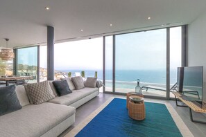 Un salon moderne avec un grand canapé, une table basse et un téléviseur. La pièce est dotée de grandes fenêtres qui offrent une vue sur l’océan.