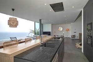 Une cuisine moderne dotée d’un grand îlot central, d’un coin repas offrant une vue sur l’océan et d’un espace salon confortable agrémenté d’un grand miroir et d’un lampadaire.