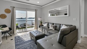 A modern living room with a grey sofa, a matching ottoman, a small side table, and a large window with a view of the sea.