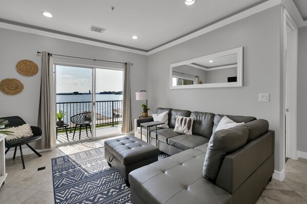 A modern living room with a grey sofa, a matching ottoman, a small side table, and a large window with a view of the sea.
