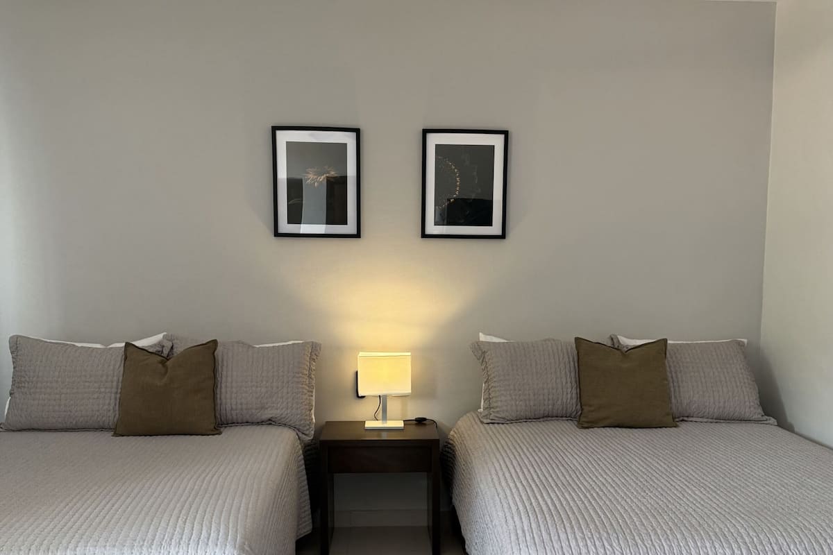 Habitación de hotel con dos camas, un ventilador de techo, una mesita de noche con lámpara y dos cuadros en la pared.