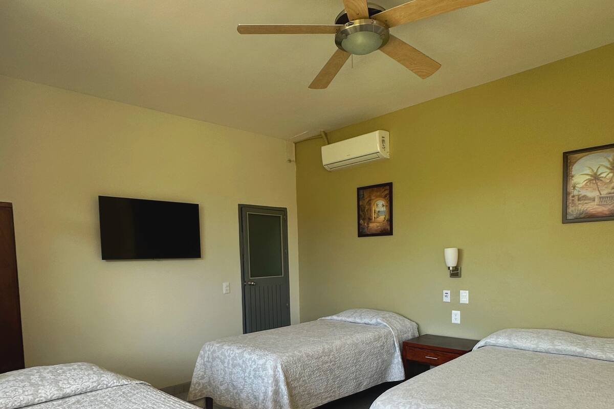 Habitación de hotel con dos camas individuales, ventilador de techo, televisor de pantalla plana, equipo de aire acondicionado y una puerta.