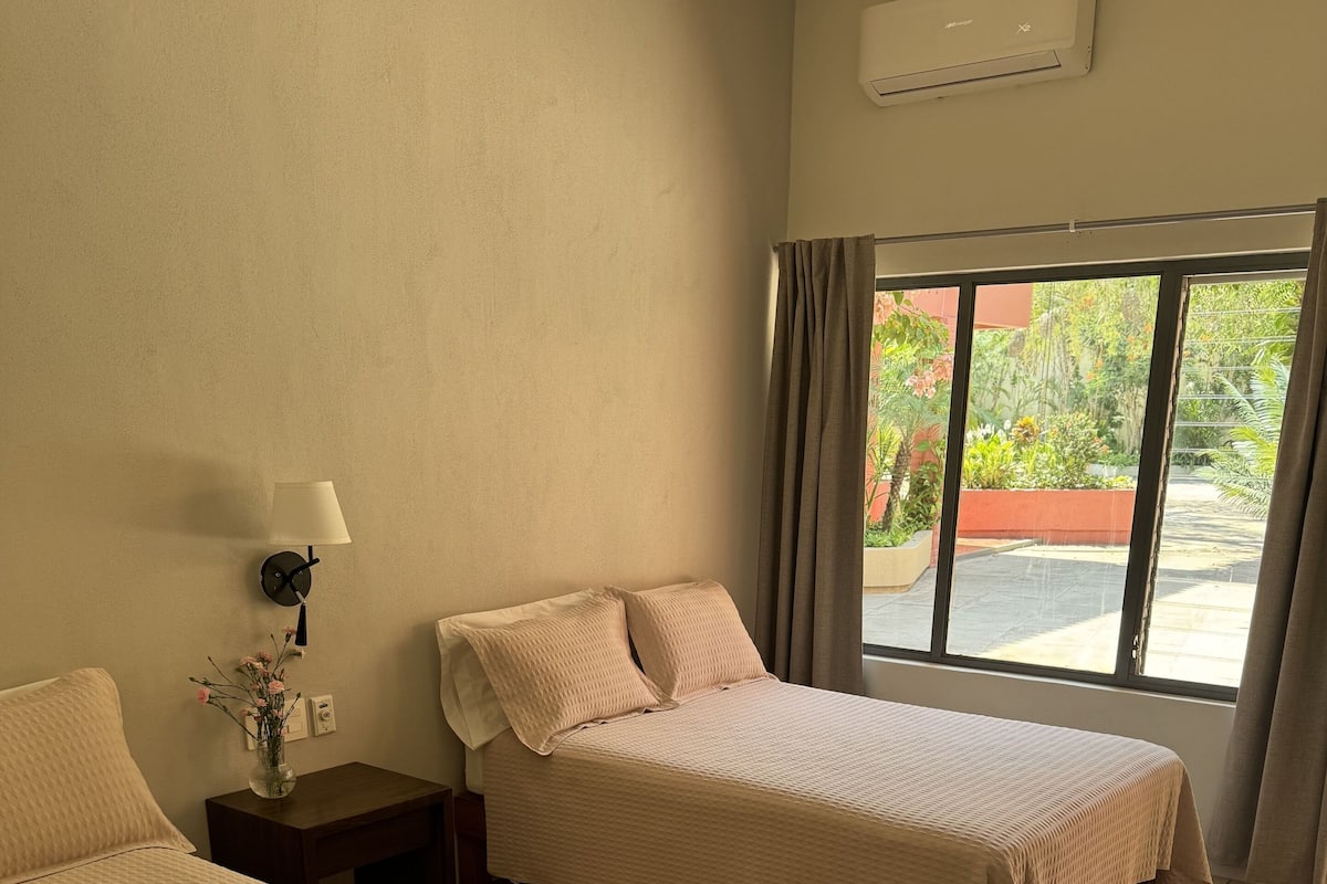 Habitación de hotel con dos camas, una mesita de noche con lámpara, una ventana con cortinas y un equipo de aire acondicionado.