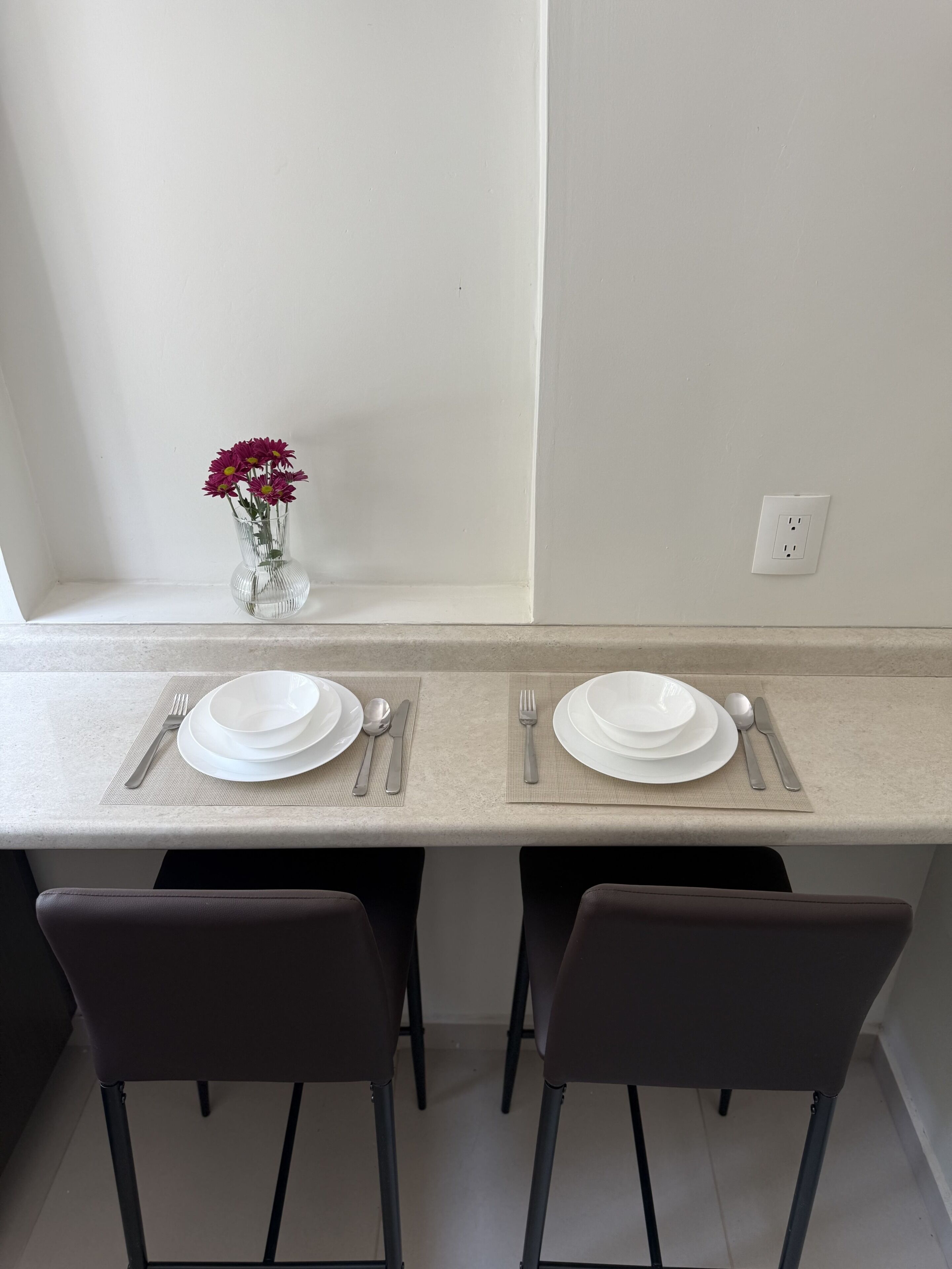 Uma área de refeições com duas cadeiras, uma mesa posta com pratos, talheres e um vaso com flores.