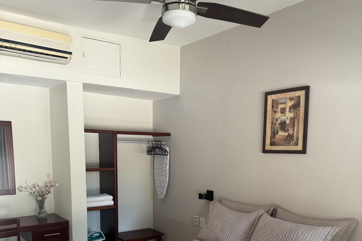 Un dormitorio con cama, ventilador de techo, un cuadro en la pared, escritorio con silla y espejo.