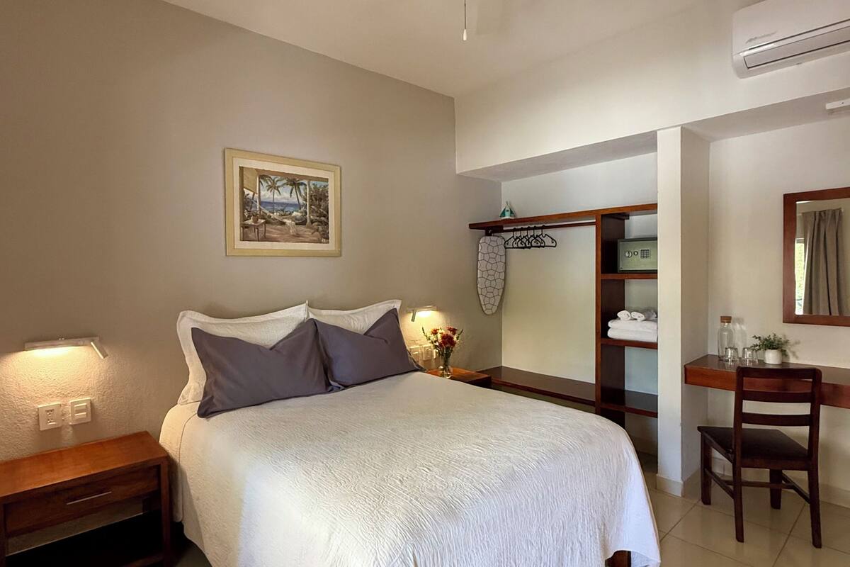 Un dormitorio con cama, mesita de noche, escritorio, silla, ventilador de techo, aire acondicionado montado en la pared y un cuadro enmarcado.