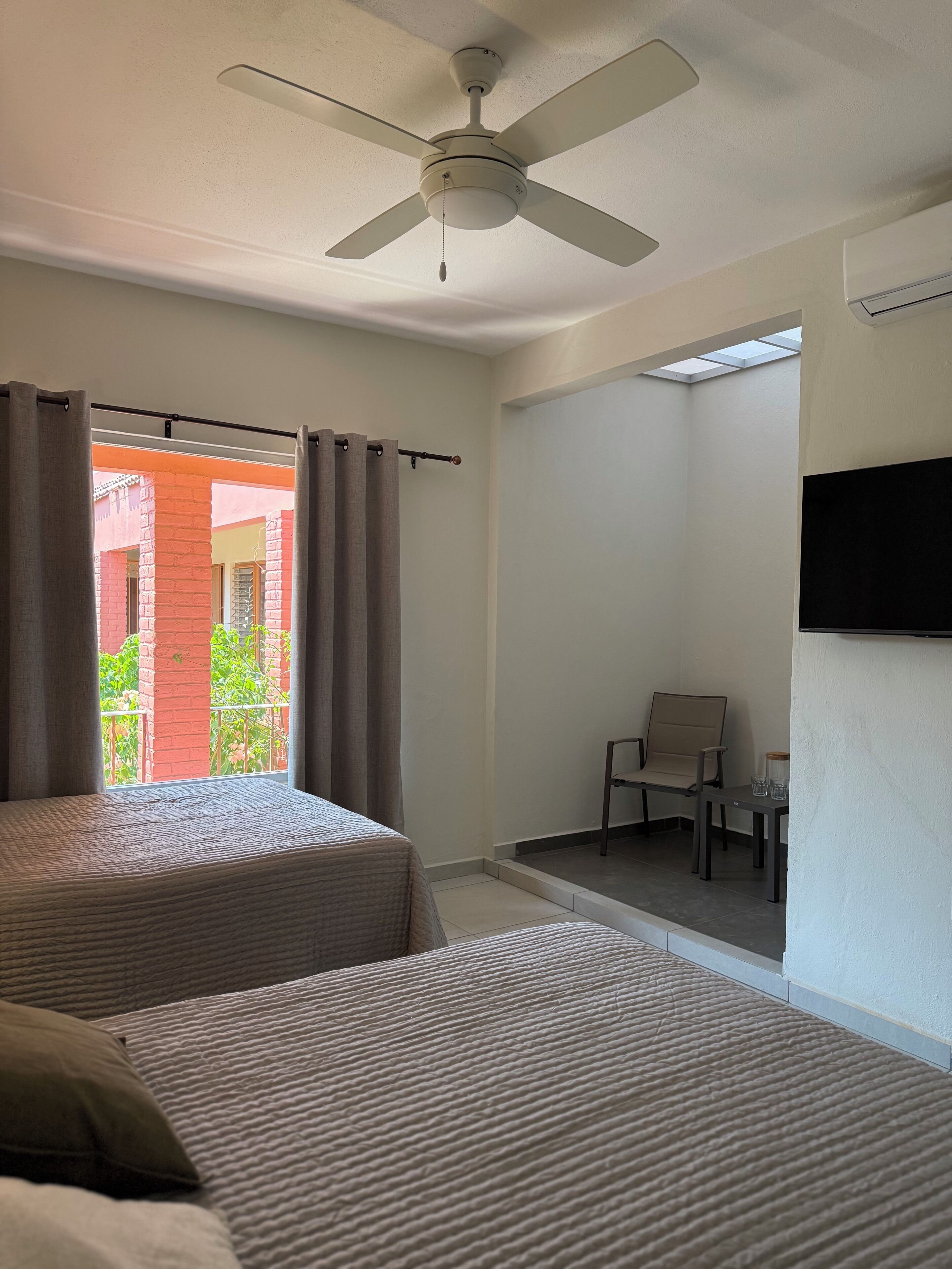 Habitación de hotel con dos camas, ventilador de techo, televisor montado en la pared y vista a un edificio de ladrillo rojo.