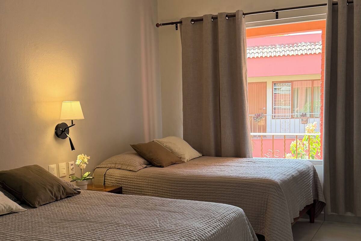 Habitación de hotel con dos camas, un ventilador de techo, una lámpara de pared y vistas a un balcón con plantas.