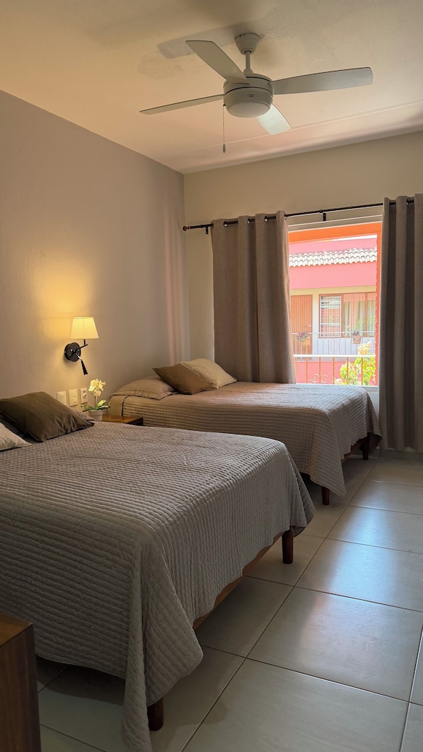 Habitación de hotel con dos camas, un ventilador de techo, una lámpara de pared y vistas a un balcón con plantas.