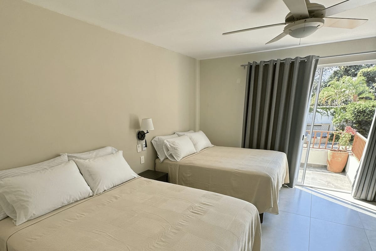 Habitación de hotel con dos camas, ventilador de techo, ventana con cortinas y vistas al exterior.