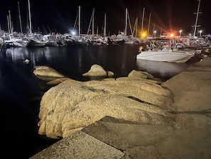 A marina at night with boats docked, a rocky breakwater, and a well-lit background.