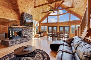 A spacious living area with a fireplace, leather sofa, wooden dining table, and large windows offering a view of the outdoors. - Stunning Views & 4-King Suites, A Dog-Friendly Cabin with Stunning Views (Sevierville)