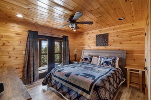 A wooden cabin bedroom with a bed, curtains, a ceiling fan, and a view through a door. - Stunning Views & 4-King Suites, A Dog-Friendly Cabin with Stunning Views (Sevierville)