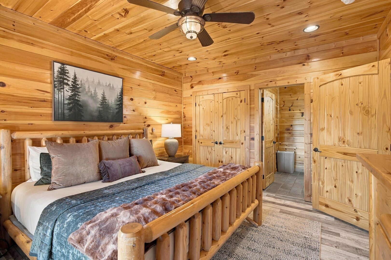 Een slaapkamer in een houten blokhut met een bed, een plafondventilator en een boslandschap op een schilderij.
