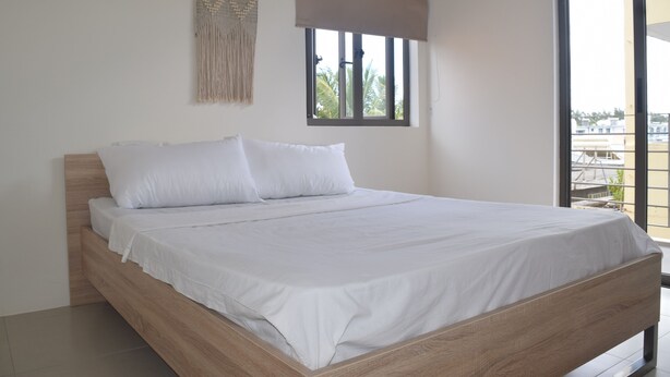 A neatly made bed with white linens in a room with a large window, a wooden headboard, and a wall hanging.