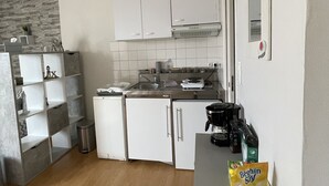 Cocina compacta con armarios blancos, estufa y cafetera. Hay estantes con utensilios de cocina y un cubo de basura.