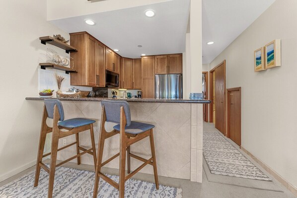 A modern kitchen with wooden cabinets, a stainless steel refrigerator, and two bar stools. - Steps to Kamaole beaches/Upgraded kitchen & baths/2 Pools/Tennis/Close to Wailea (Kihei)