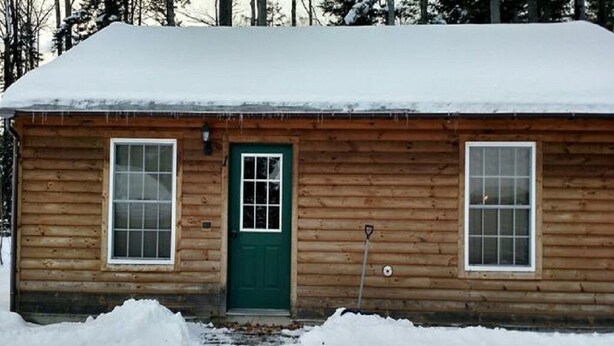 A wooden cabin with a green door, two windows, and a snow-covered roof.