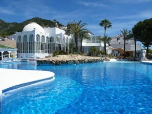 A swimming pool with a white deck, surrounded by palm trees and a white building with arched windows. - 1184 Casa el Cielo (Nerja)