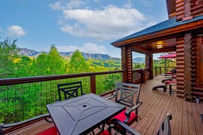 Um deck de madeira com mesa e cadeiras, com vista para uma cadeia montanhosa arborizada.