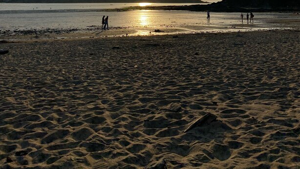 Une plage où l’on voit des gens se promener et se tenir debout, un ciel dégagé et le soleil se couchant sur l’eau.