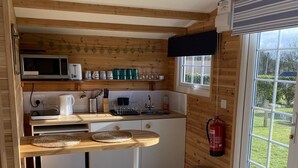 Une cuisine compacte avec des poutres en bois, un four à micro-ondes, un grille-pain et un évier. On y trouve deux tabourets et une fenêtre donnant sur un paysage verdoyant.