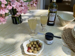 Une table recouverte d’une nappe à rayures, une bouteille de pastis, deux verres contenant une boisson jaune, un bol, une assiette d’olives et un pot de tapenade.