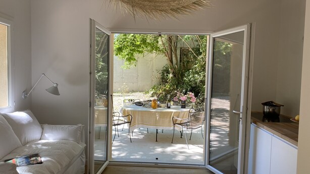 Une pièce dotée d’une porte-fenêtre coulissante donnant sur un patio extérieur. À l’intérieur, on trouve un canapé blanc, une table avec des chaises et un coin cuisine avec des placards.