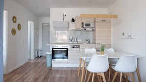 A modern kitchen with white cabinetry, a built-in oven, a microwave, and a coffee maker. There is a dining area with a white table and chairs. - Modern apartment near the beach with beach chair (Grömitz)