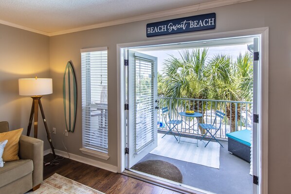 A room with a door leading to a balcony with a view of palm trees and a sign that reads 'BEACH Sweet BEACH'.