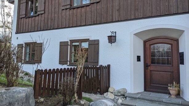A two-story building with a white exterior and brown wooden siding. It features a wooden picket fence, a stone pathway leading to a wooden door, and a small garden area with rocks and a tree.