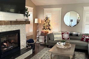 Ein gemütliches Wohnzimmer mit Kamin, Weihnachtsbaum, einer Couch mit Zierkissen und einem runden Spiegel an der Wand.