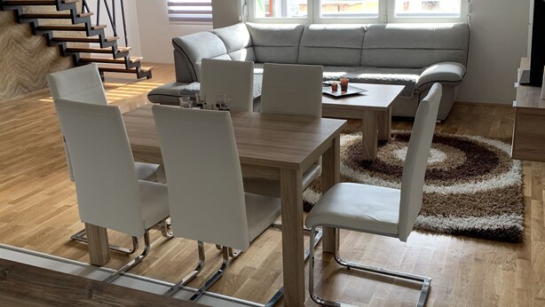 Un salon moderne avec un escalier en bois, un canapé gris, une table à manger avec des chaises et de grandes fenêtres équipées de stores.