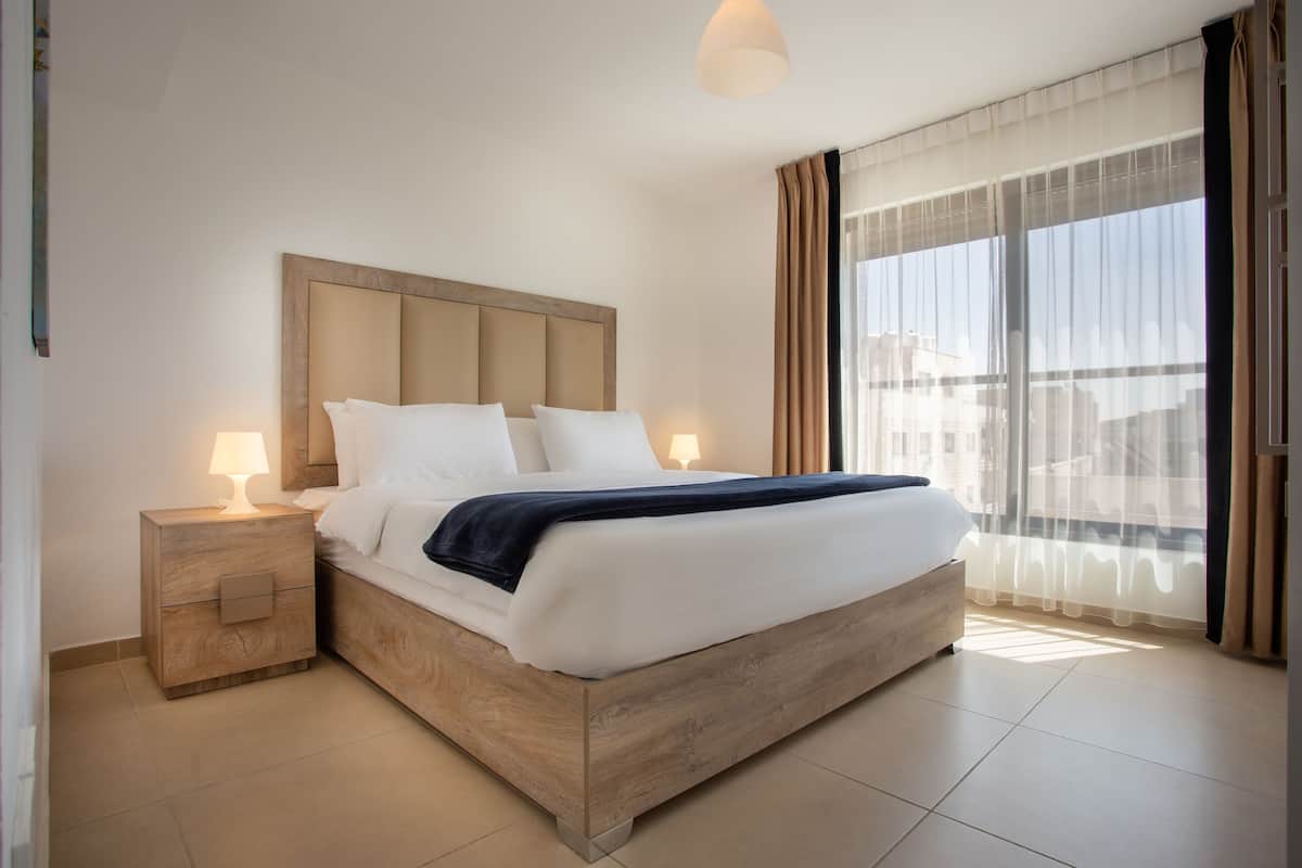 A hotel room with a large bed, two bedside tables with lamps, a wooden headboard, and a view through sheer curtains.